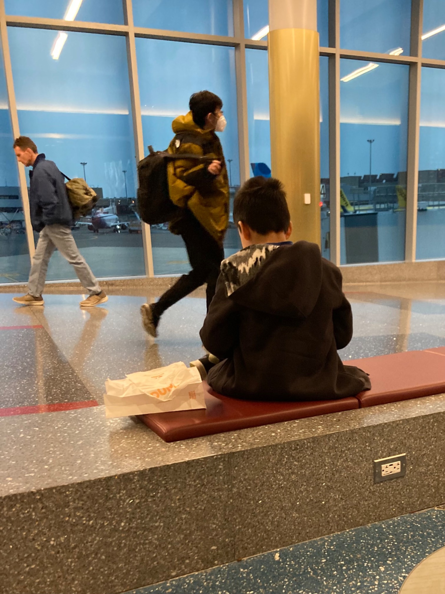 People walking by a kid sitting on a bench at the airport