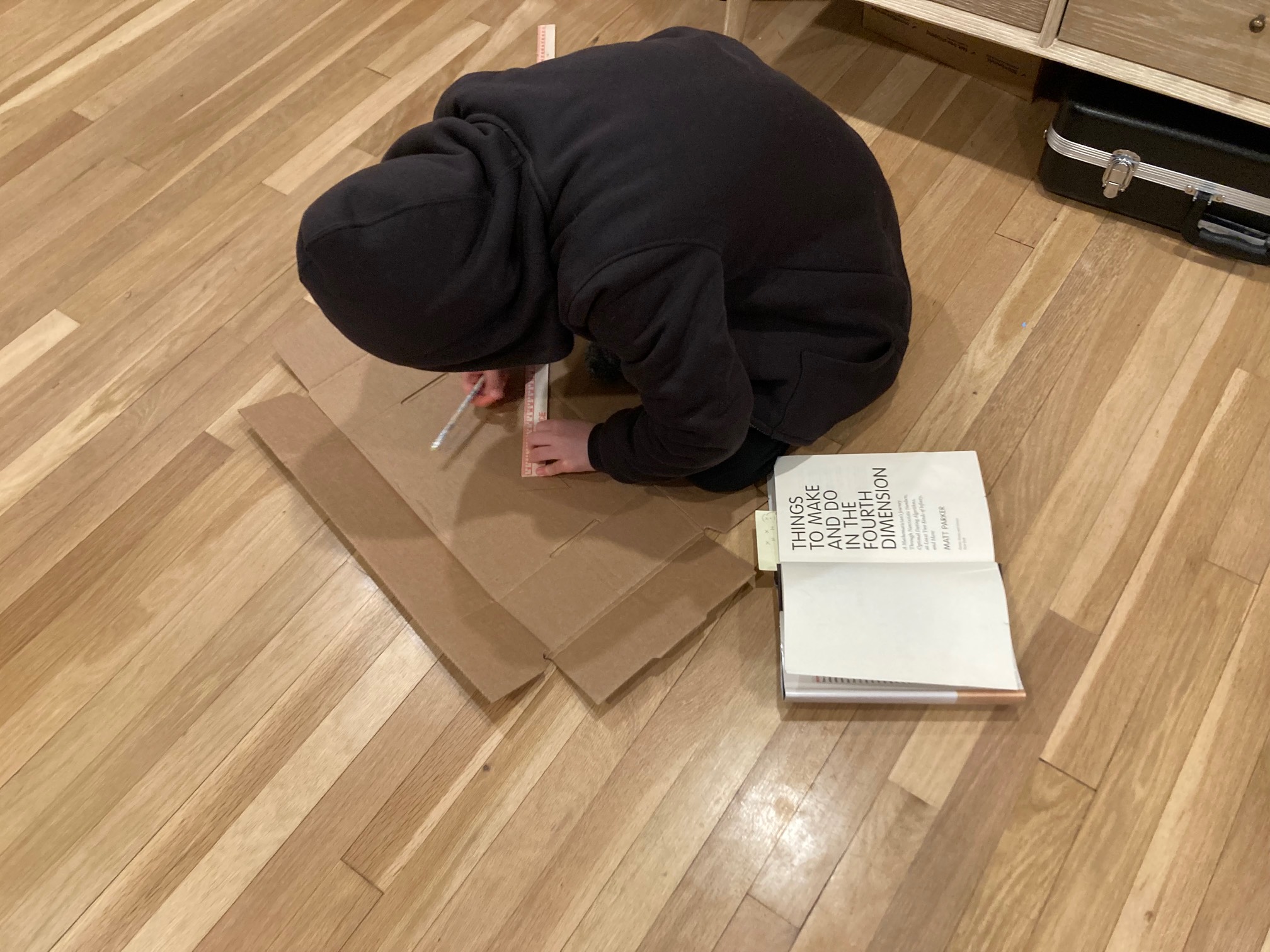 Kid crouched over cardboard and a pencil next to the book Things to Make and Do in the Fourth Dimension